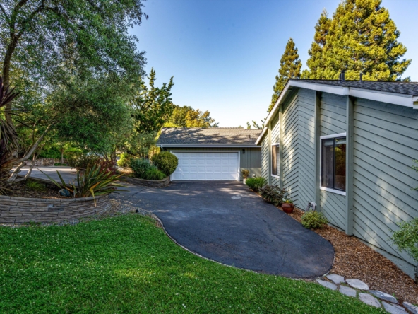 Front yard of a green-siding house with a curved asphalt driveway and a white two-car garage, surrounded by trees.