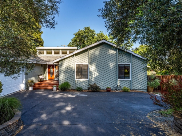 Front view of a light-blue house with a red front door, white garage, and a wooden porch, framed by leafy trees and blue sky.