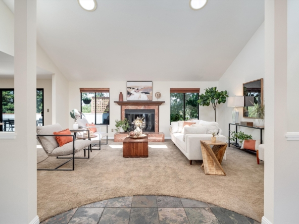Bright living room with beige carpet, white sofa, and a marble fireplace, surrounded by plants and large windows at the back.