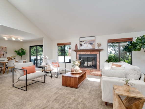 Open-concept living room with beige carpet, light furnishings, wood fireplace, and large windows letting in natural light.