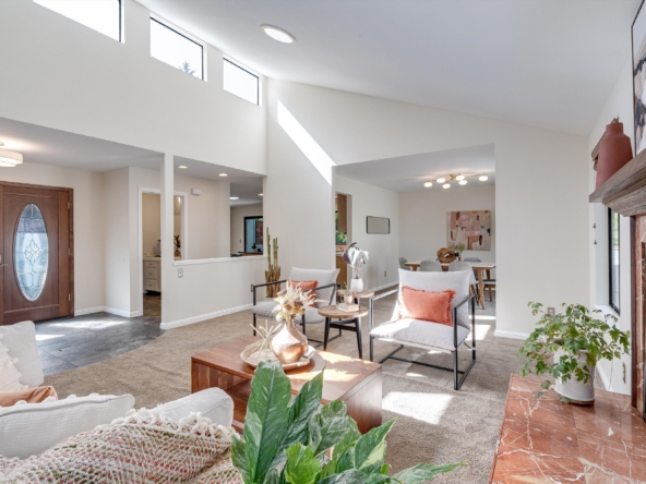 Bright modern living room with high vaulted ceiling, large windows, and a marble-tiled fireplace on the right, with cozy seating and plants.",