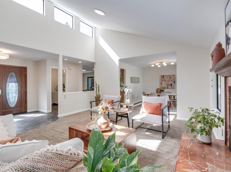 Bright modern living room with high vaulted ceiling, large windows, and a marble-tiled fireplace on the right, with cozy seating and plants.",