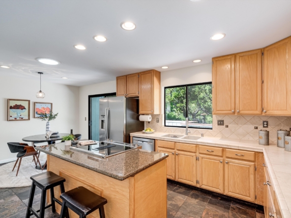 Bright kitchen with maple cabinets, granite island, and stainless appliances beneath a large window overlooking trees.