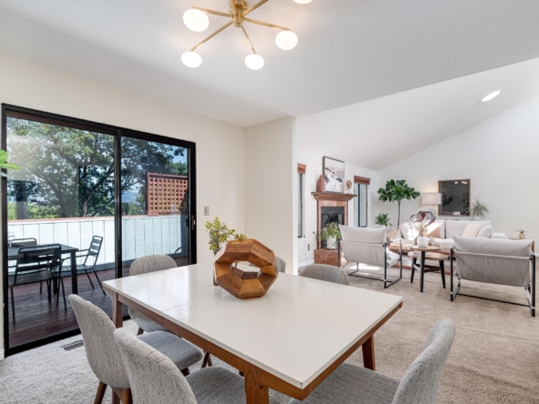 Open-concept dining area with white table and gray chairs, sliding glass door to outdoor deck, and living room beyond with fireplace and plants.
