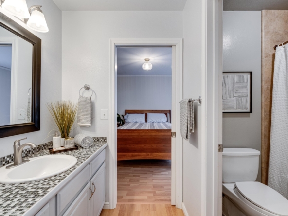 Bathroom vanity with mosaic countertop and sink on the left; doorway opens to a lit bedroom with a wooden headboard beyond.