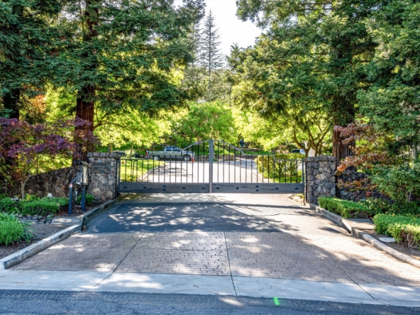 Gated stone entrance to a leafy park, with a brick-paved road leading through the gate and trees on both sides of the driveway.