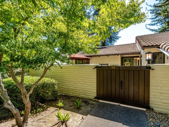Front yard view of a beige house with a dark brown gate, flanked by lush green trees and a tiled roof.