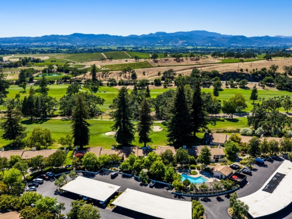Aerial view of a golf course with green fairways, trees, and a surrounding residential area, with hills in the distance under a blue sky.
