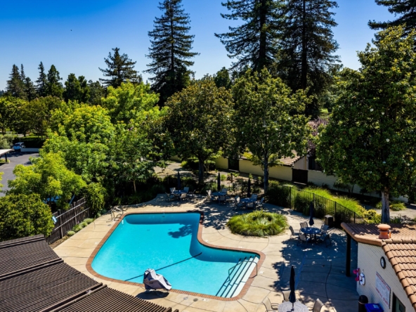 Outdoor swimming pool area with blue water, lounge chairs, and tables, bordered by trees under a clear blue sky