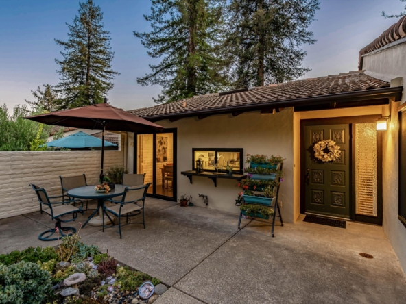 Patio seating area with a round table and metal chairs, two umbrellas, and a line of potted plants by the house door.