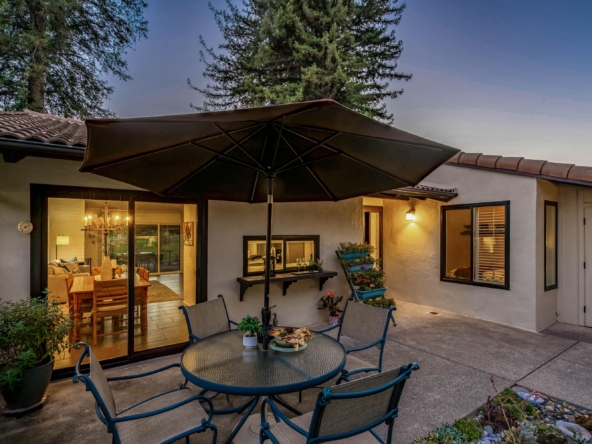 Backyard patio with a large umbrella over a glass table and chairs, adjacent to a house with warm interior lights.