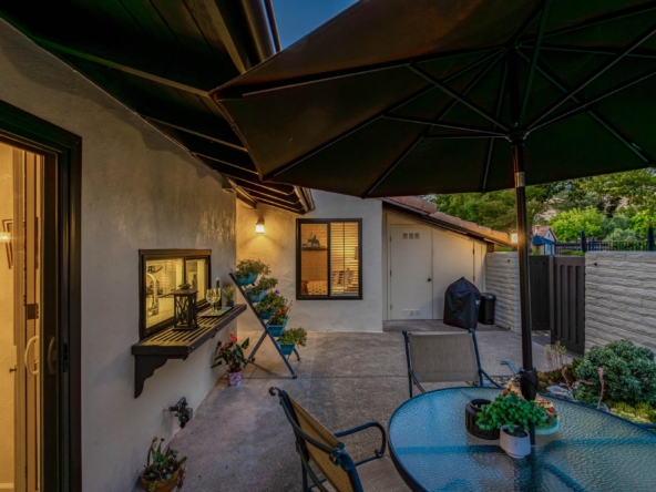 Backyard patio at dusk with a large black umbrella, glass table and metal chairs, potted plants, and exterior house lighting
