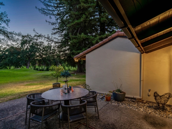 Outdoor patio scene at dusk with a round table and several chairs near a stucco house and wooden eaves, trees and lawn beyond.
