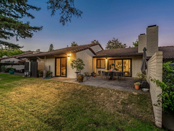 Evening view of a suburban backyard patio: a single-story house with a tiled roof, sliding doors, outdoor table and chairs, and warm exterior lights.