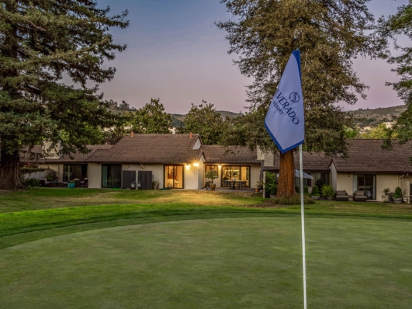 Flag on a pole beside a resort lawn, with a white flag and blue trim reading 'Resort' in front of a row of houses and tall trees at dusk.