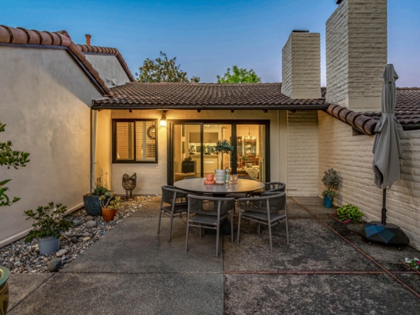 Outdoor backyard patio scene with a round dining table and six chairs set on a concrete slab, plants in pots, and a warm-lit interior visible through sliding glass doors; blue evening sky above and stone chimneys in the background.