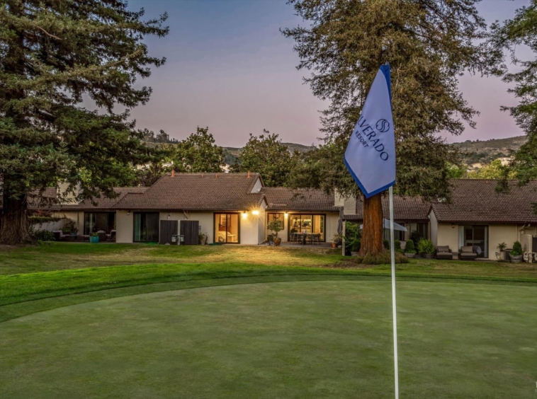 Putting green with a flagstick in the foreground and a clubhouse lit at dusk in the background, surrounded by trees and manicured grass.